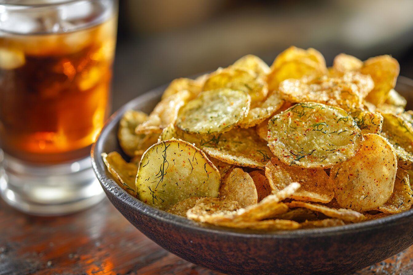 Bowl of crispy pickle chips on a wooden table with iced tea.